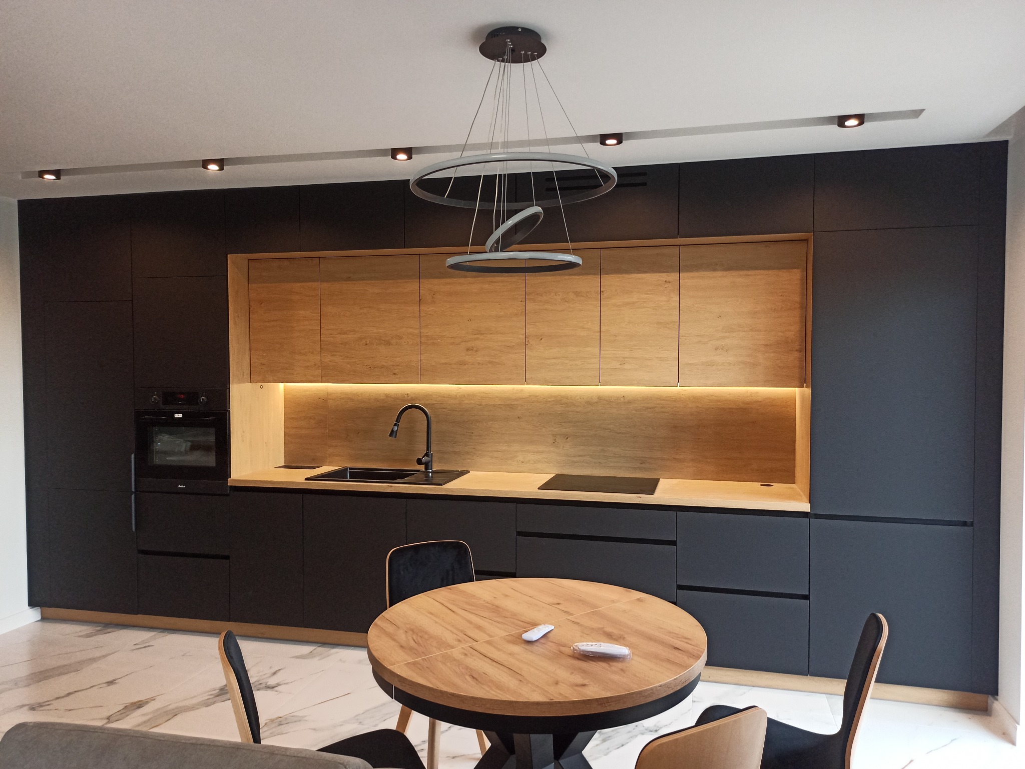 Nowoczesna kuchnia: czarne matowe fronty, drewniane akcenty, okrągły stół z drewnianym blatem, designerska lampa. Minimalizm i elegancja w detalu.