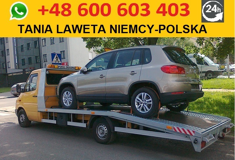 Żółta laweta z beżowym Volkswagenem Tiguan zabezpieczonym pasami transportowymi, zaparkowana na ulicy z widocznym znakiem drogowym i budynkiem w tle.