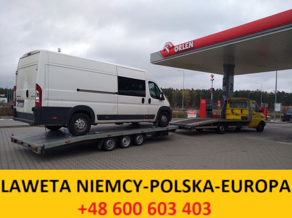 Biały bus na szarej platformie lawety, zaparkowany przy stacji benzynowej Orlen, z żółtą lawetą z tyłu.