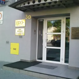 Elewacja budynku z szarą, chropowatą fakturą, z białą stolarką okienną i drzwiową, tabliczkami informacyjnymi firm i oznaczeniem gazu, zabezpieczona przed graffiti.