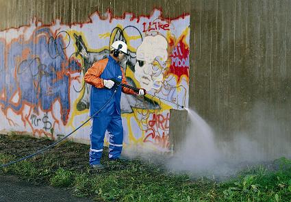 Pracownik w kombinezonie ochronnym usuwa graffiti z betonowej ściany za pomocą myjki ciśnieniowej.