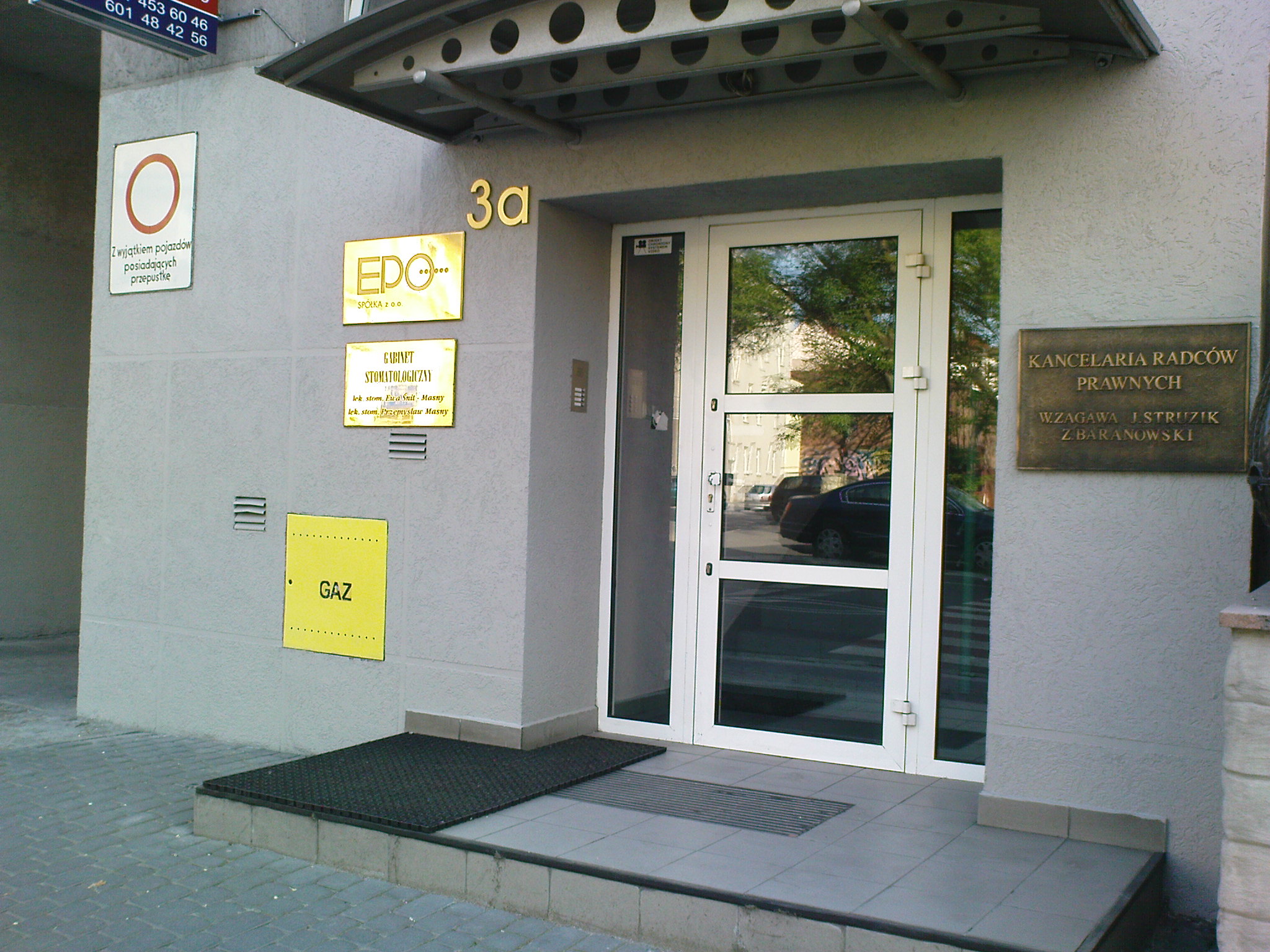 Elewacja budynku z szarą, chropowatą fakturą, z białą stolarką okienną i drzwiową, tabliczkami informacyjnymi firm i oznaczeniem gazu, zabezpieczona przed graffiti.