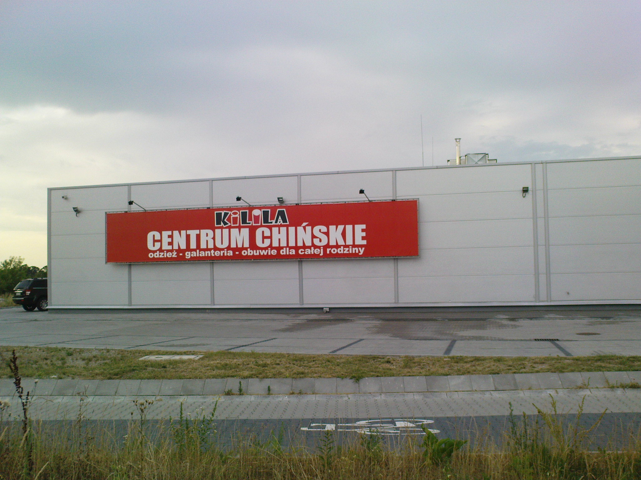 Duży, pomalowany na biało budynek z zamontowanym, szerokim, czerwonym banerem reklamowym z napisem 'KiLOLA CENTRUM CHIŃSKIE odzież - galanteria - obuwie dla całej rodziny'. Baner oświetlony...