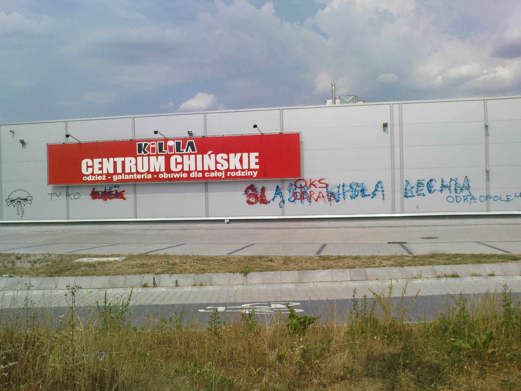 Biała ściana budynku z czerwonym szyldem 'KILILA CENTRUM CHIŃSKIE' i graffiti, widoczne zabezpieczenia anty-graffiti.
