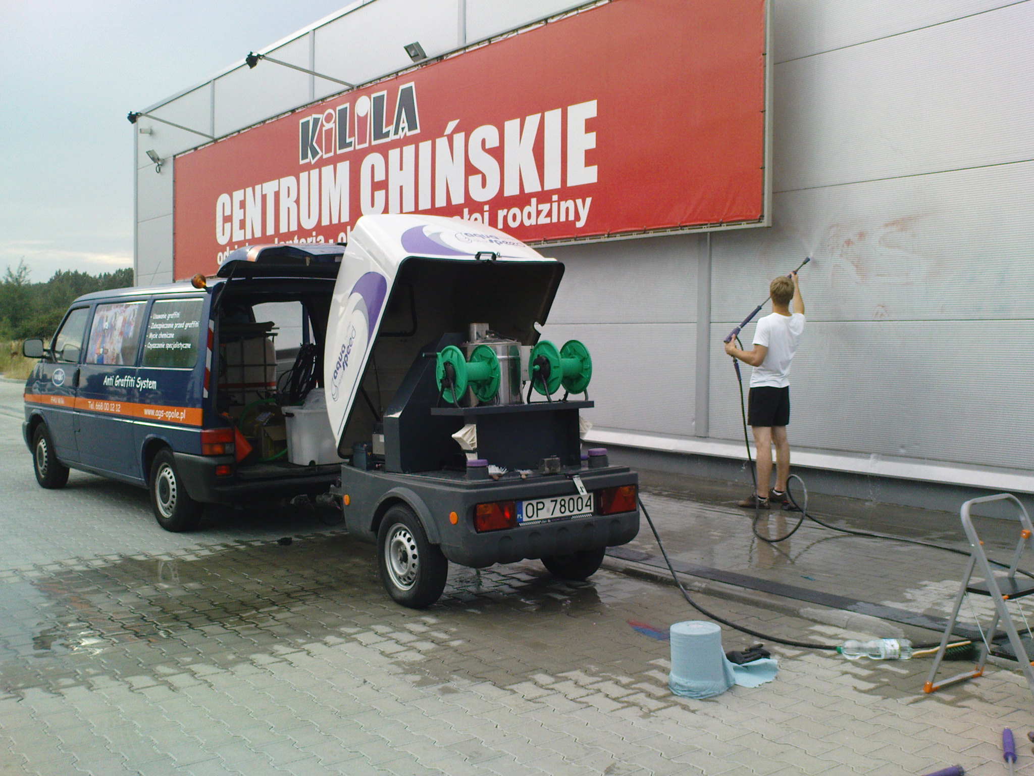 Pracownik w krótkich spodenkach usuwa graffiti z elewacji budynku handlowego za pomocą myjki ciśnieniowej; na pierwszym planie zaparkowany bus z przyczepką wyposażoną w sprzęt do usuwania graffiti.