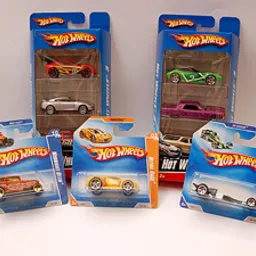 Zestaw zapakowanych samochodzików Hot Wheels w różnorodnych kolorach i modelach, niektóre w opakowaniach zbiorczych po trzy sztuki, inne pojedynczo, ustawione na jasnym tle.