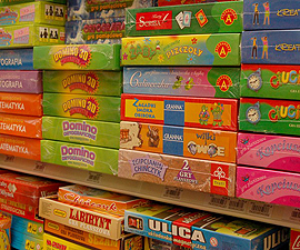 Półka sklepowa wypełniona różnorodnymi grami planszowymi i logicznymi w kolorowych opakowaniach, ustawionymi jedna na drugiej, widoczne tytuły gier takie jak Domino, Labirynt, Ulice, Wilki i inne.