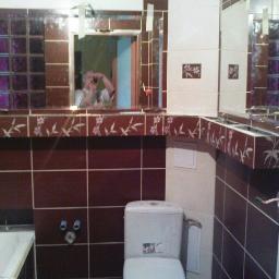 Usługi Remontowo-Wykończeniowe - Łazienka w trakcie prac wykończeniowych z brązowo-białymi płytkami, widoczne przyłącza hydrauliczne i toaleta, w lustrze odbija się osoba robiąca zdjęcie.