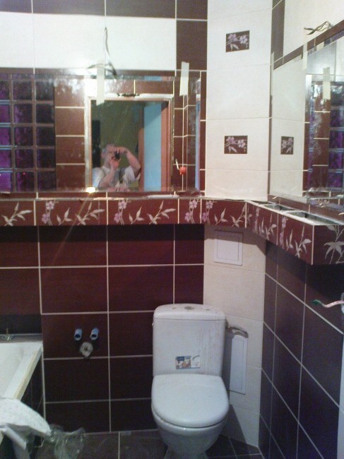 Łazienka w trakcie prac wykończeniowych z brązowo-białymi płytkami, widoczne przyłącza hydrauliczne i toaleta, w lustrze odbija się osoba robiąca zdjęcie.