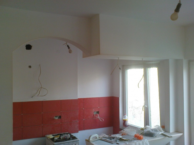 Wnętrze kuchni w trakcie prac wykończeniowych: ściany częściowo pokryte czerwoną glazurą, świeżo zagruntowana górna część ścian przygotowana do malowania, widoczne wystające przewody elektryczne...