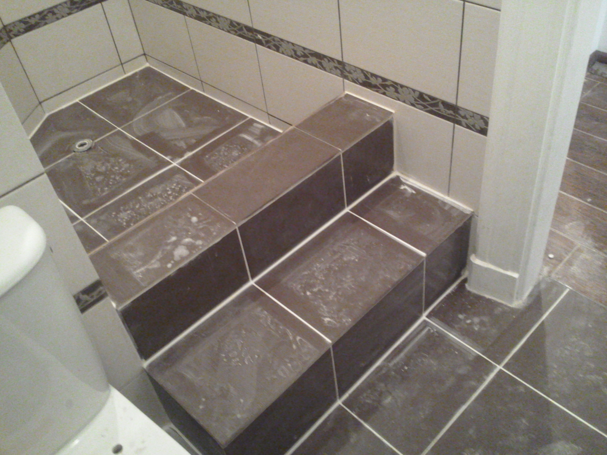 Wykończona łazienka z dwustopniowym podestem wyłożonym brązowymi płytkami, widoczny odpływ liniowy, fragment toalety i dekoracyjny pas płytek na ścianie.