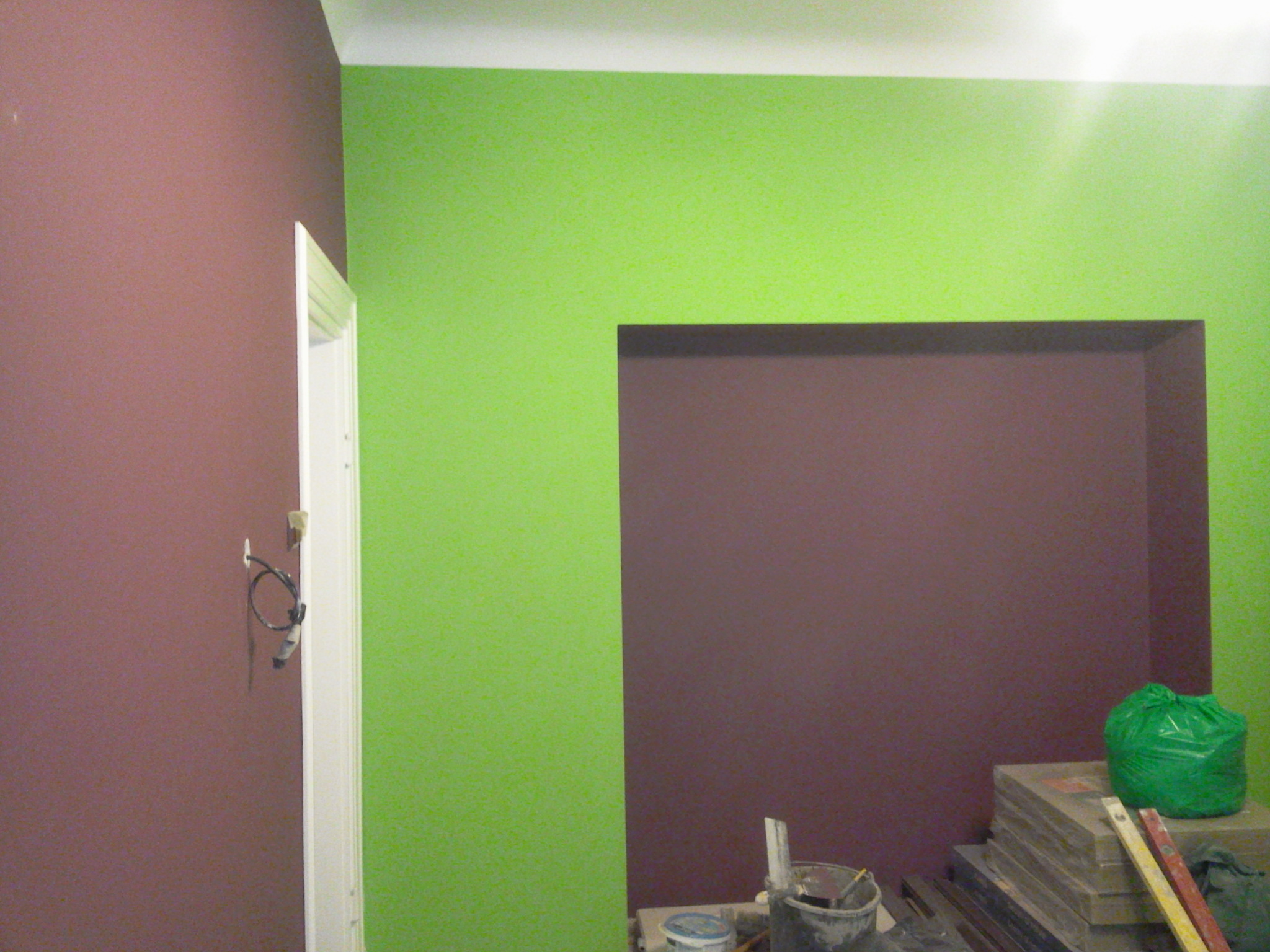 Ściana pomalowana na dwa kolory: intensywny zielony i stonowany fiolet, z widocznym otworem w ścianie również w kolorze fioletowym. Przy ścianie narzędzia malarskie i wiadro z farbą.