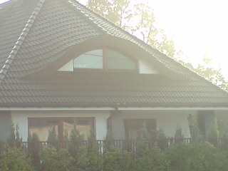 Nowo wybudowany dom jednorodzinny w Ząbkach z ciemną dachówką i białymi ścianami, widoczny z poziomu ogrodu. Uwagę zwraca półokrągłe okno na poddaszu.