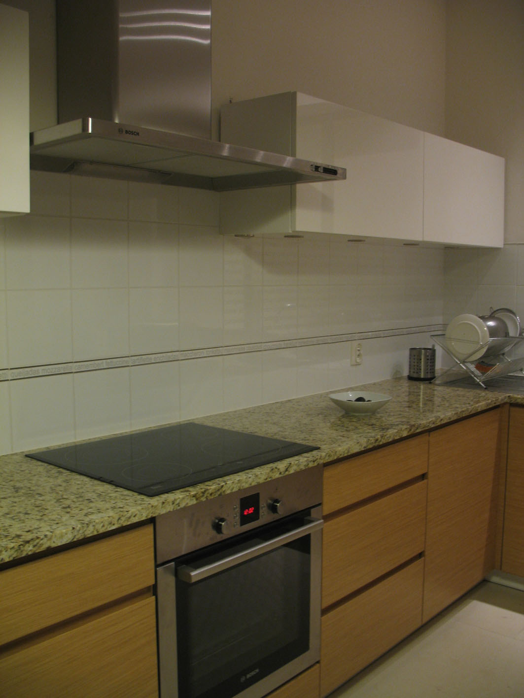 Nowoczesna kuchnia z beżowymi, drewnopodobnymi szafkami, blatem z granitu, płytą indukcyjną, piekarnikiem marki Bosch i okapem ze stali nierdzewnej.