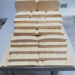 Stos jasnych, drewnianych desek podłogowych z różnymi profilami, ułożonych na stole, widoczne detale połączeń i tekstura drewna.