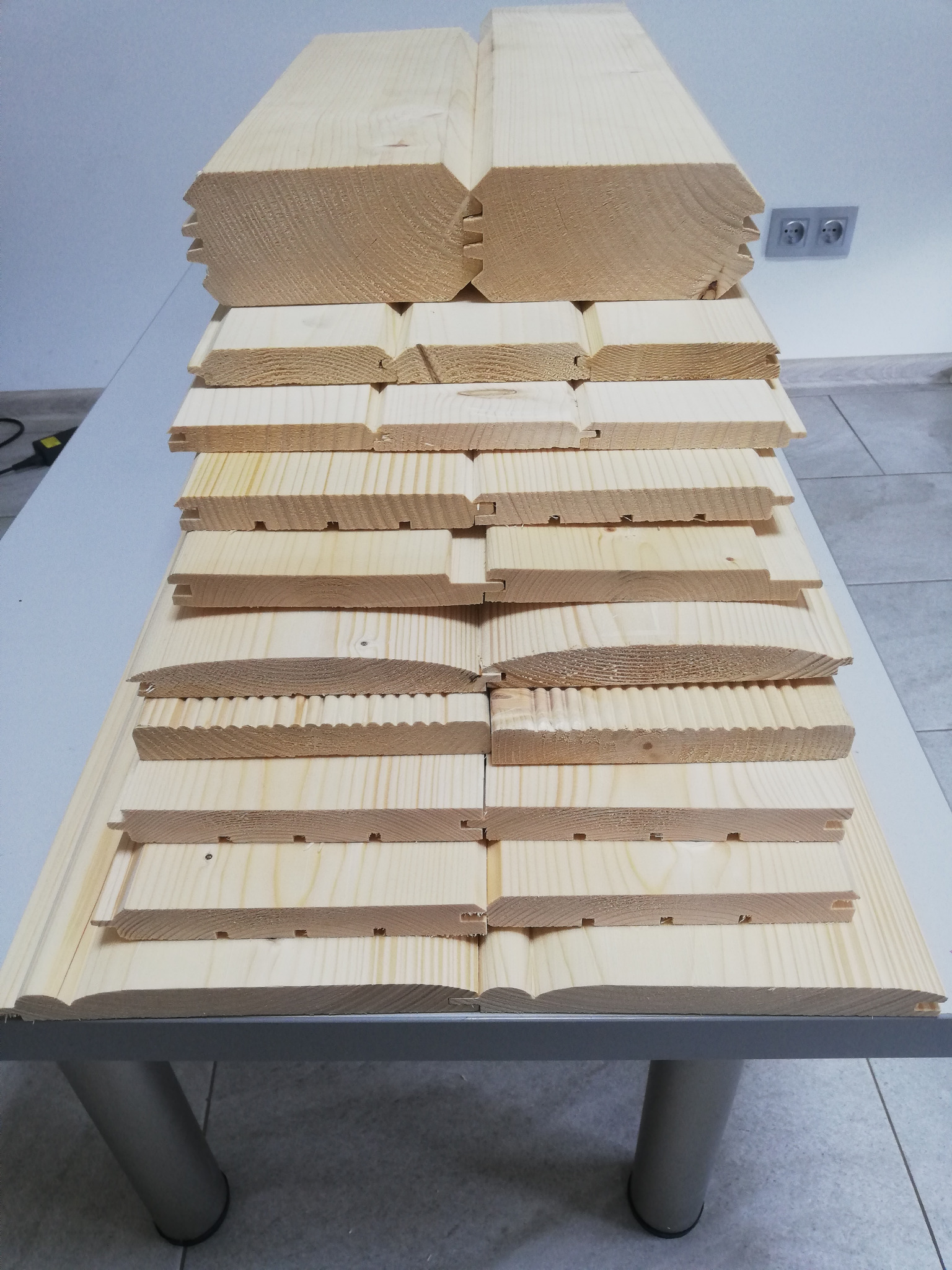 Stos jasnych, drewnianych desek podłogowych z różnymi profilami, ułożonych na stole, widoczne detale połączeń i tekstura drewna.