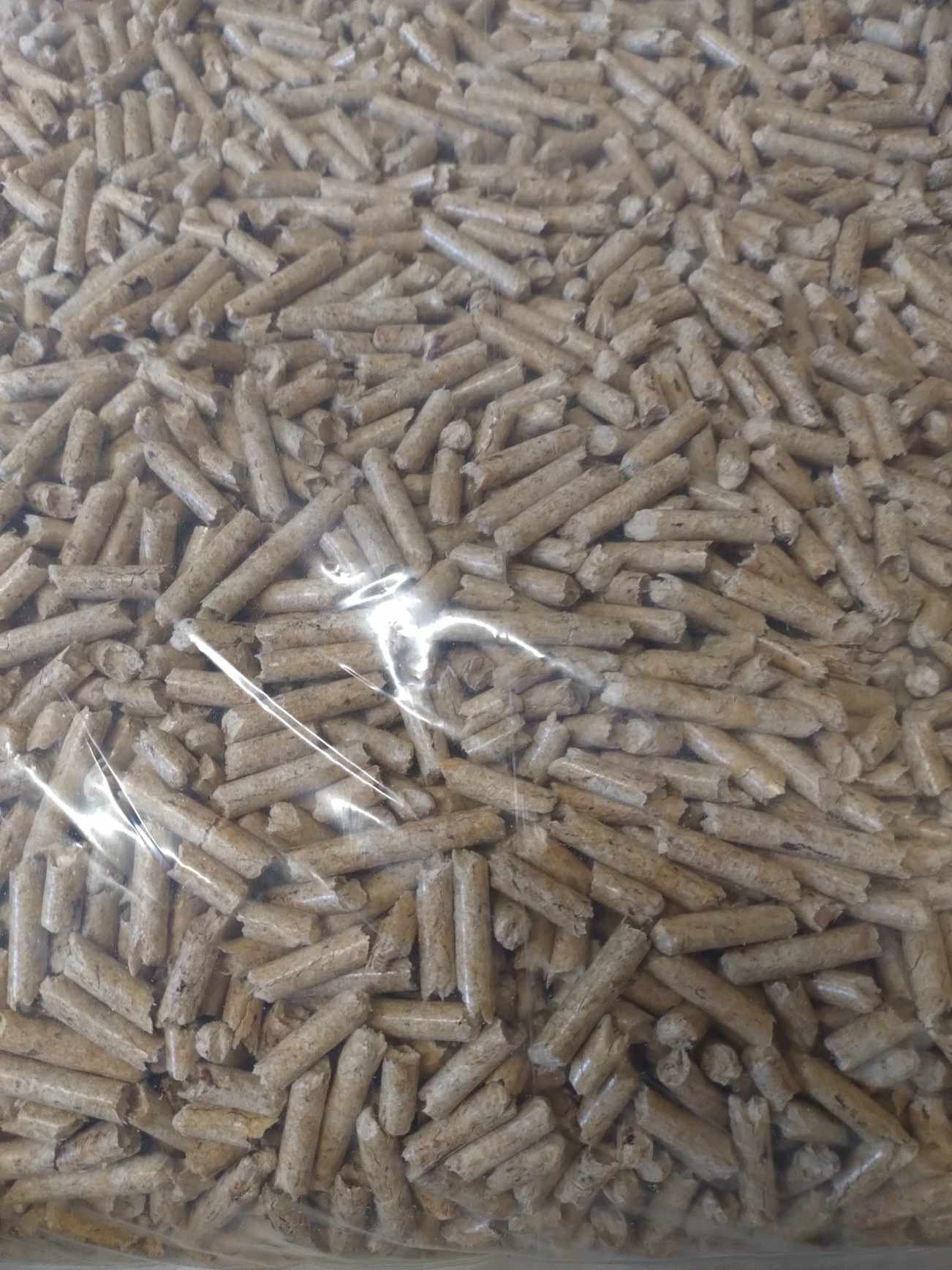 Beżowe granulki pelletu drzewnego widoczne przez przezroczyste opakowanie foliowe, z widocznymi refleksami światła.