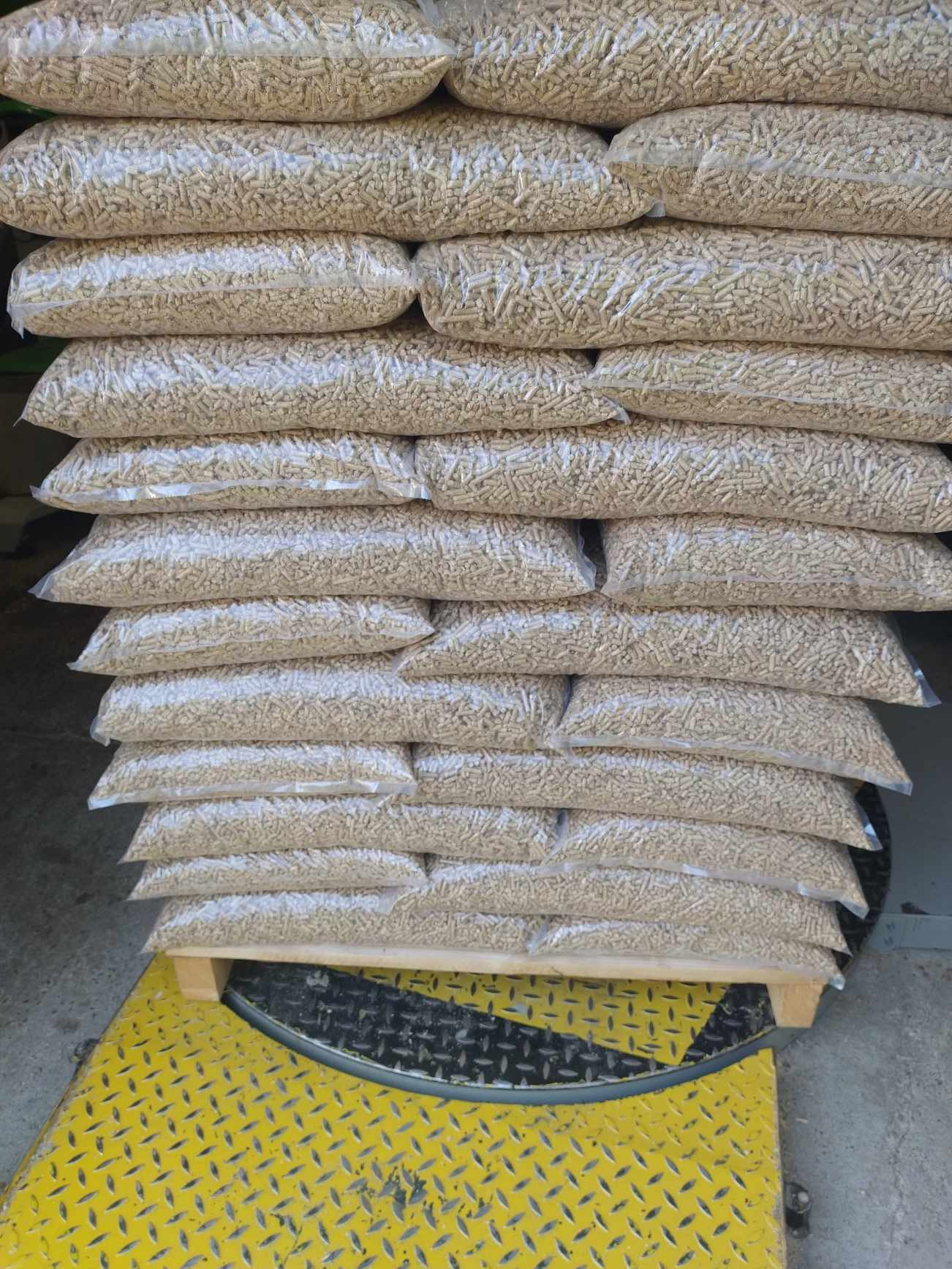 Stos przezroczystych worków wypełnionych pelletem drzewnym ułożonych na palecie z żółto-czarną, wzorzystą powierzchnią antypoślizgową.