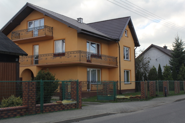 Żółty dom jednorodzinny z ciemnobrązowym dachem i ozdobnymi balkonami z kutego żelaza, otoczony ceglanym ogrodzeniem z zieloną siatką.