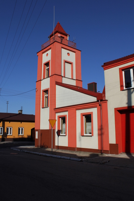 Budynek o pomarańczowo-białej fasadzie z wieżą, widoczny znak drogowy ustąp pierwszeństwa, na dachu czerwona dachówka.