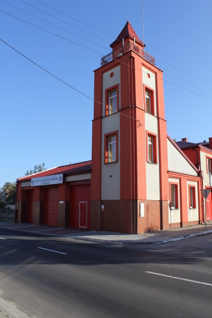 Budynek straży pożarnej w kolorach czerwieni i bieli z wieżą obserwacyjną, widoczny z poziomu ulicy w słoneczny dzień.