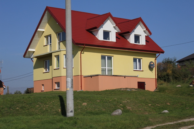 Dwupiętrowy dom z czerwonym dachem dwuspadowym i oknami z białymi ramami, pomalowany na żółto z pomarańczową podstawą, widziany z dołu pod lekkim kątem.