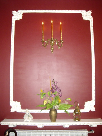 Gipsowa sztukateria ramy na bordowej ścianie, pod nią półka z wazonem kwiatów, świecznikiem i figurką, nad półką świecznik ścienny z trzema zapalonymi świecami.