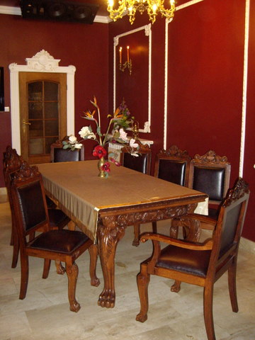 Jadalnia z ciemnoczerwonymi ścianami, ozdobną sztukaterią, drewnianym stołem z rzeźbionymi nogami w kształcie łap i krzesłami z ciemną tapicerką.