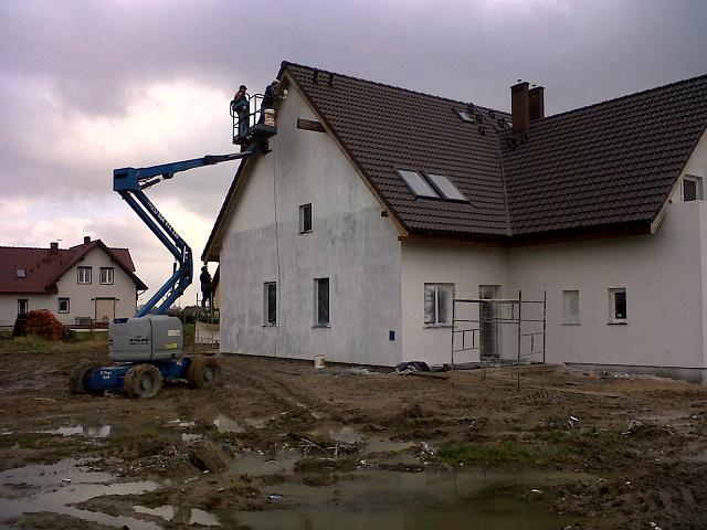 Prace elewacyjne na wysokości wykonywane z użyciem podnośnika koszowego Genie na tle zachmurzonego nieba, widoczna ocieplona ściana budynku w trakcie malowania.