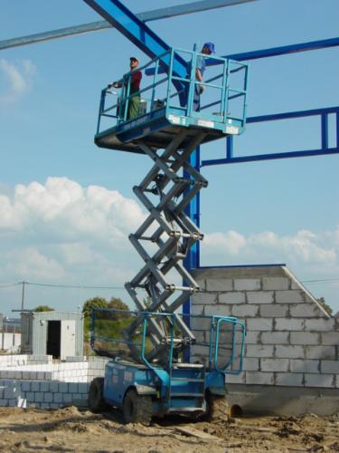 Dwóch pracowników na podnośniku nożycowym przy montażu stalowej konstrukcji hali, widoczne bloczki betonowe i kontener w tle.