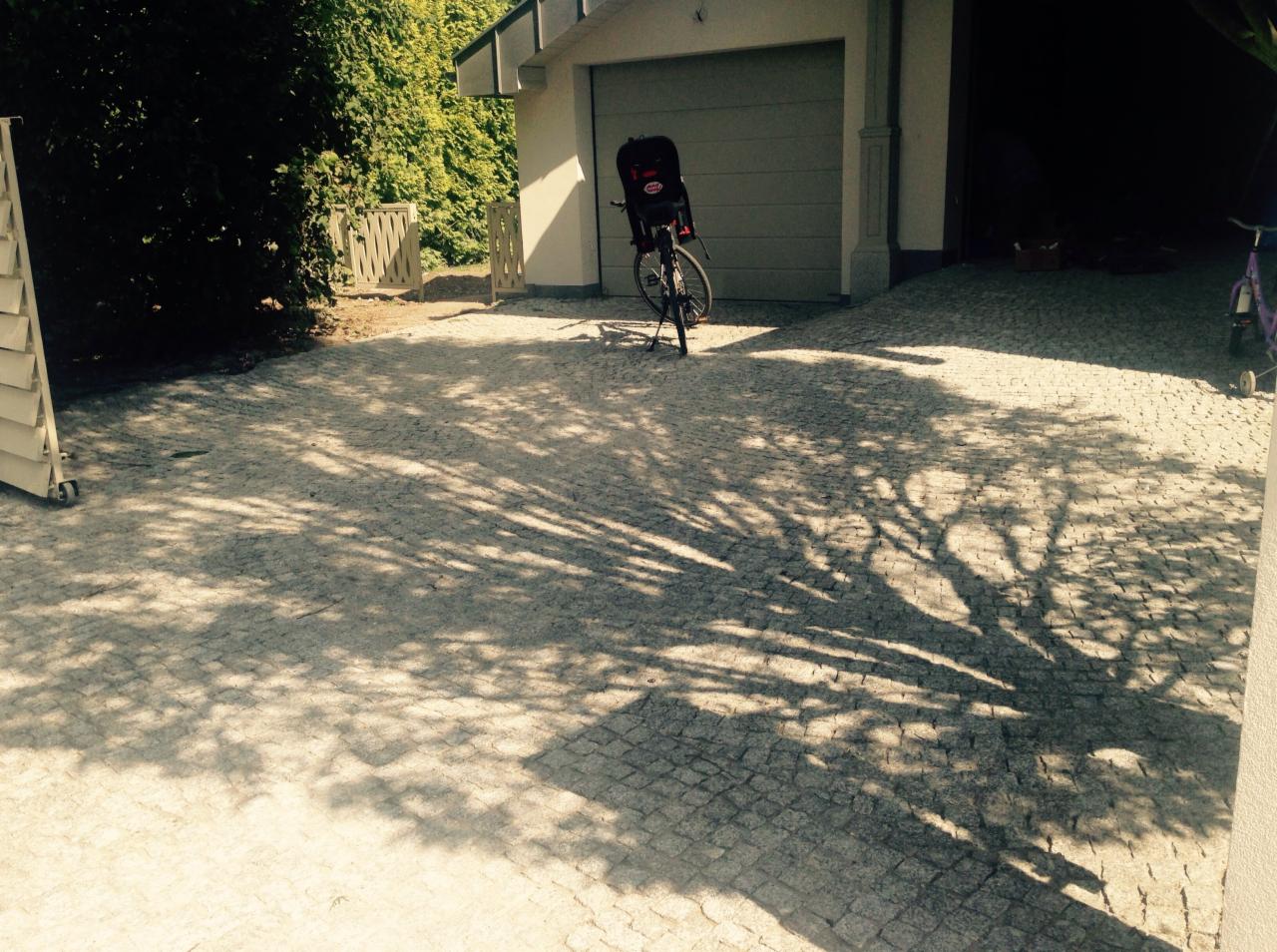 Granitowa kostka brukowa na podjeździe w cieniu drzewa, z rowerem z fotelikiem dziecięcym i otwartym garażem w tle.