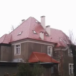Budynek z nowym, czerwonym dachem z dachówki ceramicznej, widoczne okna dachowe i kominy, szara elewacja budynku, pochmurne niebo w tle.