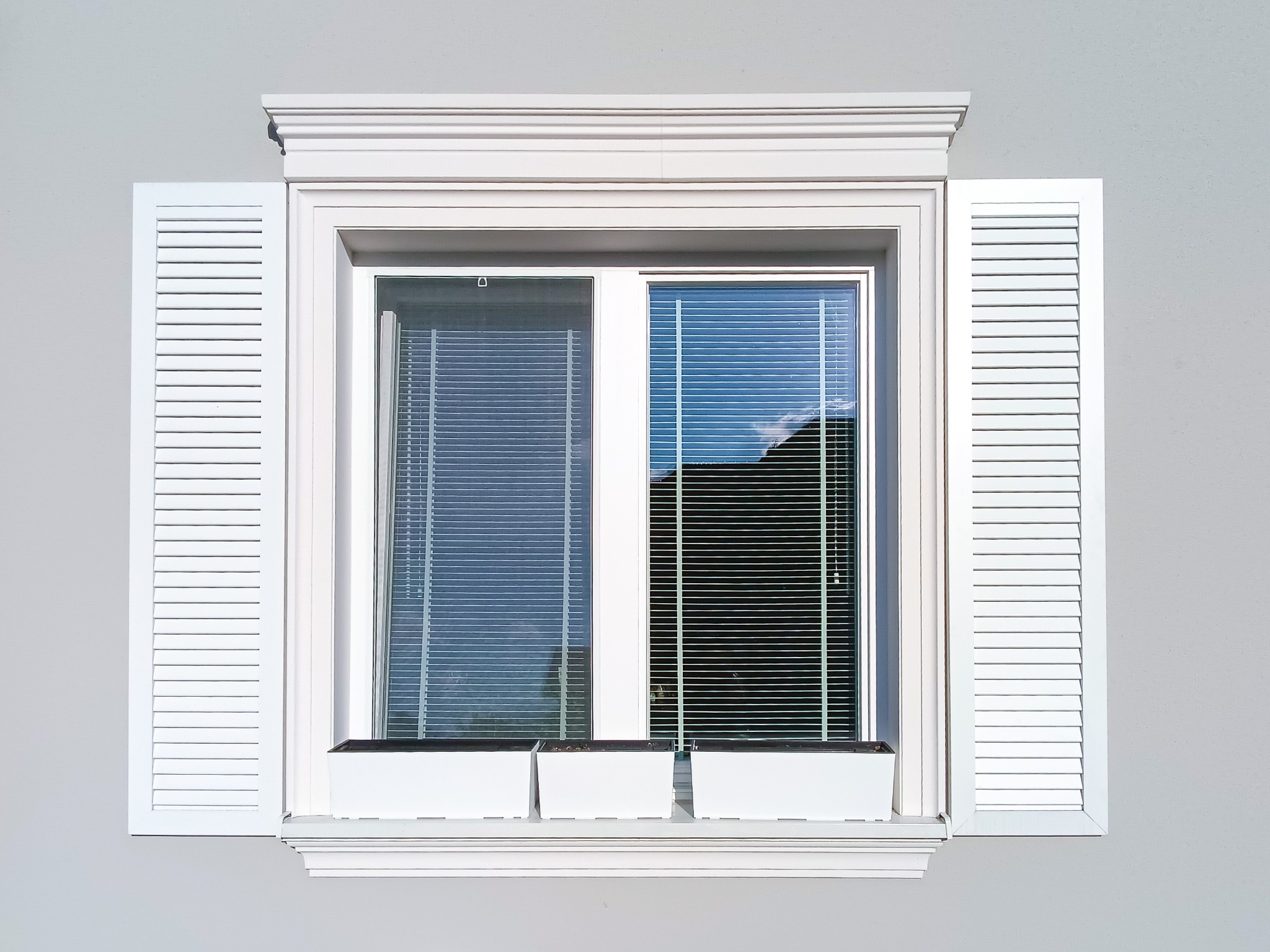 Białe okno z louverowymi okiennicami i sztukaterią okienną, nadproże i opaski boczne w klasycznym stylu. W oknach żaluzje. Pod oknem trzy białe donice.