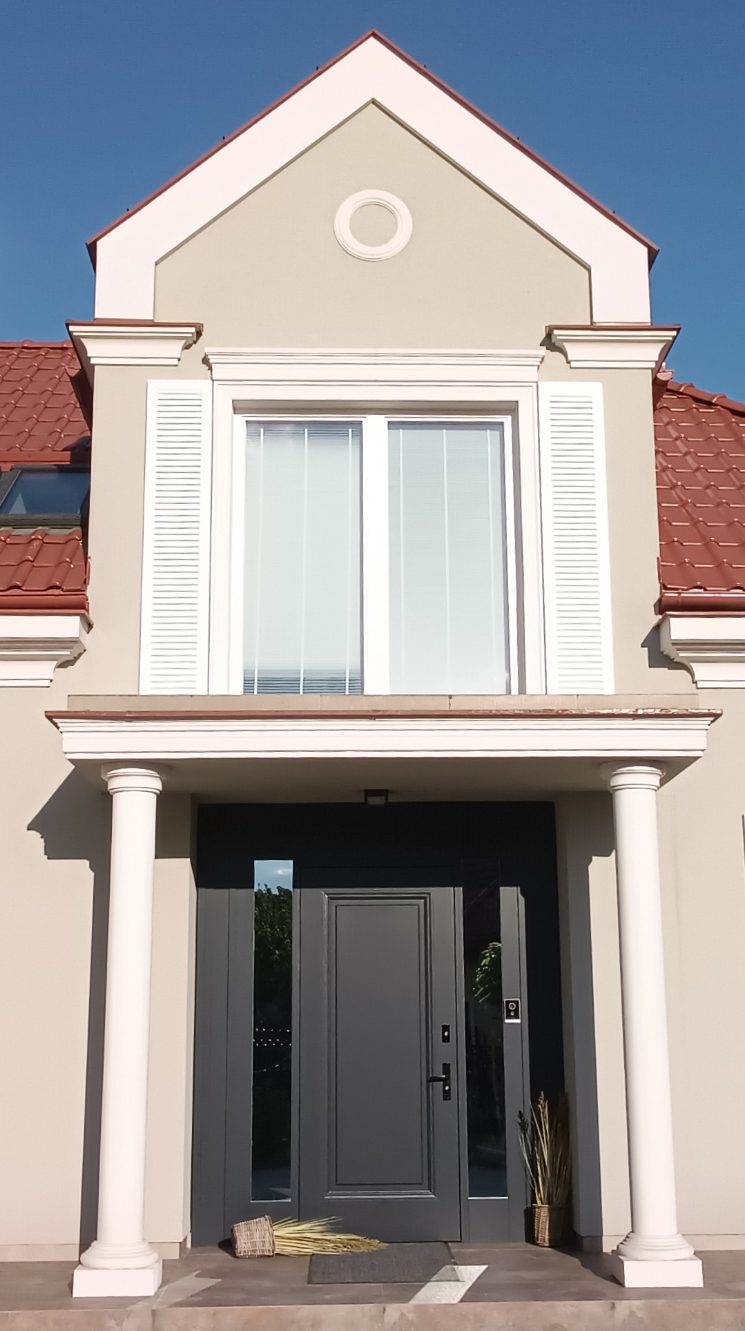 Elewacja domu jednorodzinnego z kolumnami, szarą stolarką okienną i drzwiami. Dachówka ceramiczna w kolorze czerwonym. Dekoracyjne elementy architektoniczne.