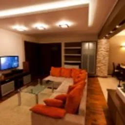 Jasne wnętrze salonu z narożną sofą w odcieniach beżu i pomarańczy, szklany stolik kawowy, ściana akcentująca z kamienia dekoracyjnego, oświetlenie sufitowe punktowe i listwowe, drewniana podłoga.