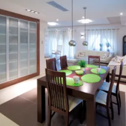 Jasne, przestronne wnętrze z drewnianym stołem jadalnym nakrytym zielonymi podkładkami, zabudowaną szafą z przesuwnymi, matowymi drzwiami i częścią wypoczynkową z białymi sofami w tle.