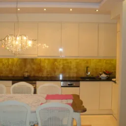 Jasna kuchnia z błyszczącymi szafkami, czarnym blatem i dekoracyjnym panelem ściennym w odcieniach brązu i złota, oświetlona kryształowym żyrandolem i punktowymi lampami sufitowymi, widok na stół...