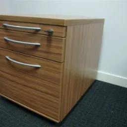 Drewniana szafka biurowa z trzema szufladami, zamykana na klucz, na tle białej ściany i ciemnej wykładziny.