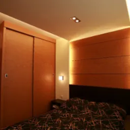 Sypialnia z drewnianą zabudową ściany i szafą przesuwną, oświetlenie punktowe i nocne lampki, widok na łóżko z wzorzystą pościelą.