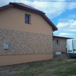 Wykończona elewacja domu z beżowym tynkiem i dekoracyjną, kamienną okładziną. Na ścianie zamontowana skrzynka elektryczna. W tle biały bus i błękitne niebo.