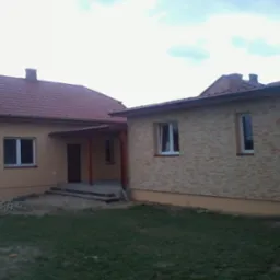 Jasny, parterowy dom z czerwonym dachem i elewacją w odcieniach beżu, częściowo wykończoną kamieniem dekoracyjnym. Widoczne okna i zadaszone wejście.