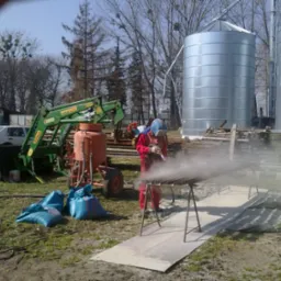 Pracownik w czerwonym kombinezonie i ochronnej masce piaskuje metalowe profile na zewnątrz, obok widoczny agregat do piaskowania, worki z piaskiem i maszyna rolnicza, w tle silosy zbożowe.