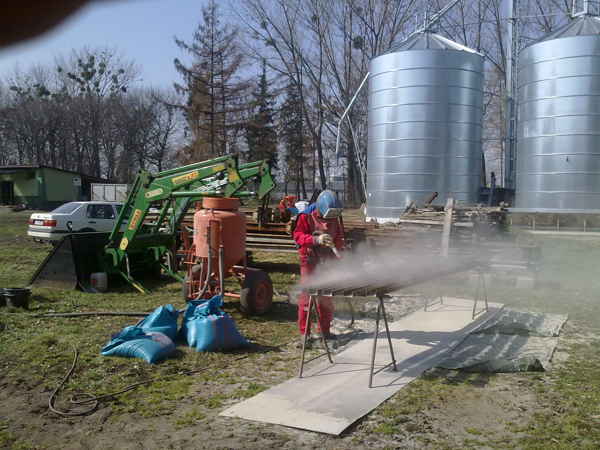 Pracownik w czerwonym kombinezonie i ochronnej masce piaskuje metalowe profile na zewnątrz, obok widoczny agregat do piaskowania, worki z piaskiem i maszyna rolnicza, w tle silosy zbożowe.