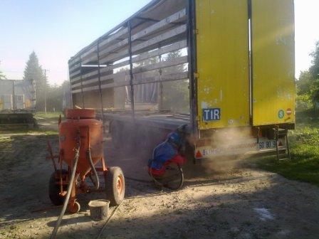 Przyczepa TIR w trakcie czyszczenia strumieniem ściernym, widoczny pracownik w kombinezonie ochronnym oraz pomarańczowy agregat do piaskowania.