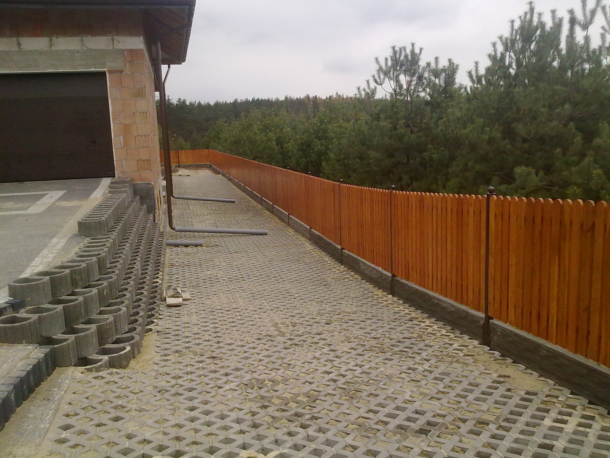 Nowe drewniane ogrodzenie panelowe z metalowymi słupkami, widok z boku na podjazd wyłożony ażurowymi płytami betonowymi, w tle budynek w trakcie budowy i las.