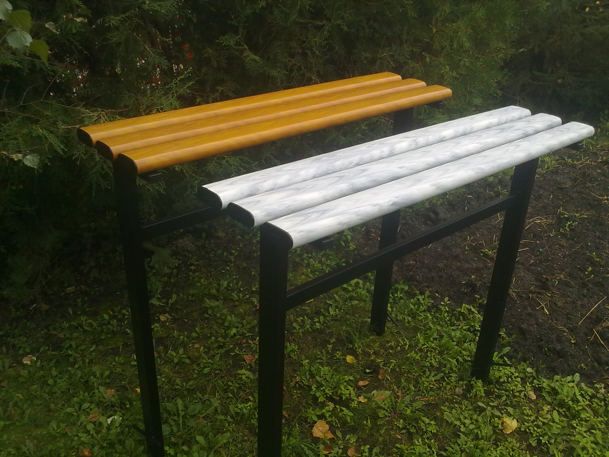 Dwie ławeczki cmentarne: jedna z drewnianym siedziskiem w kolorze miodowym, druga z siedziskiem imitującym marmur, obie na czarnych metalowych nogach, na tle zieleni.