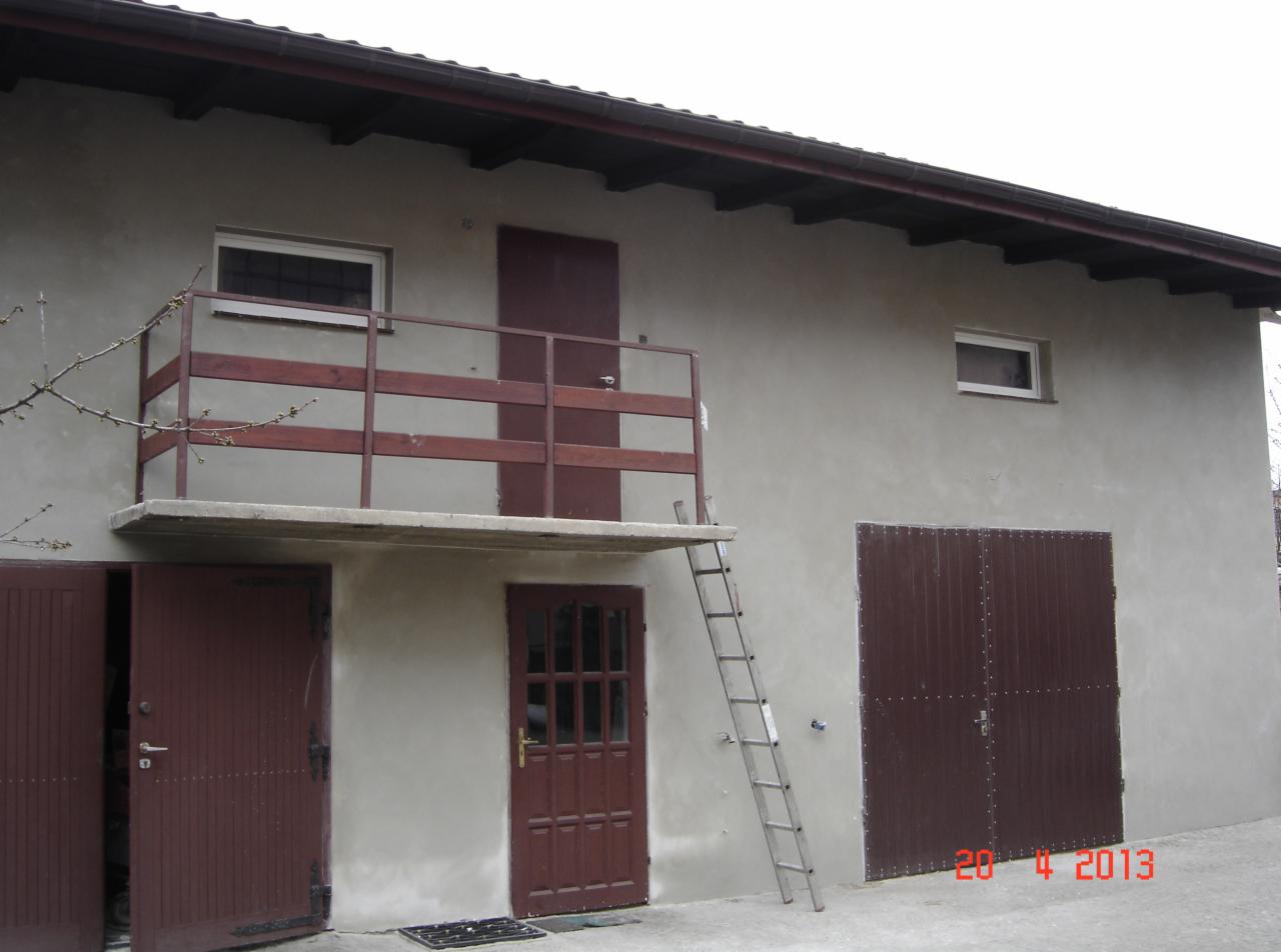 Budynek gospodarczy z balkonem i drabina, szare tynki, brązowe drzwi i brama garażowa. Widoczny fragment drzewa i data na dole zdjęcia.