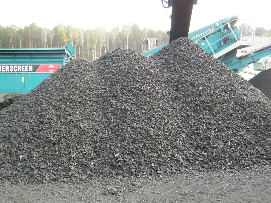 Góra węgla kamiennego o frakcji groszku, usypana na placu składowym, z widocznymi przesiewaczami i sortownikami w tle.