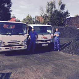 Dwa białe samochody dostawcze Isuzu i TwojOpał na tle hałdy węgla. Dwóch mężczyzn w niebieskich kombinezonach. Transport i sprzedaż opału.