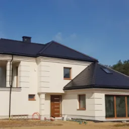 Nowoczesny dom jednorodzinny z antracytowym dachem karpiówkowym i jasną elewacją, widoczne panele słoneczne w tle.